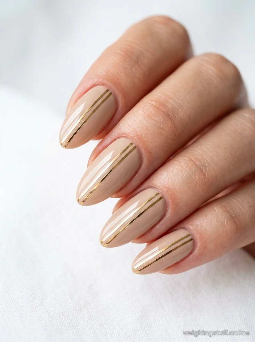 Close-up of manicured nails with beige polish and thin gold metallic stripes on a white background, showcasing minimalist nail art.